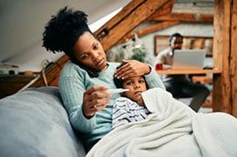 Mom, on the phone, cuddles son while holding a thermometer.