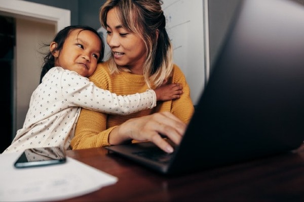 Mom uses laptop while daughter hugs her.